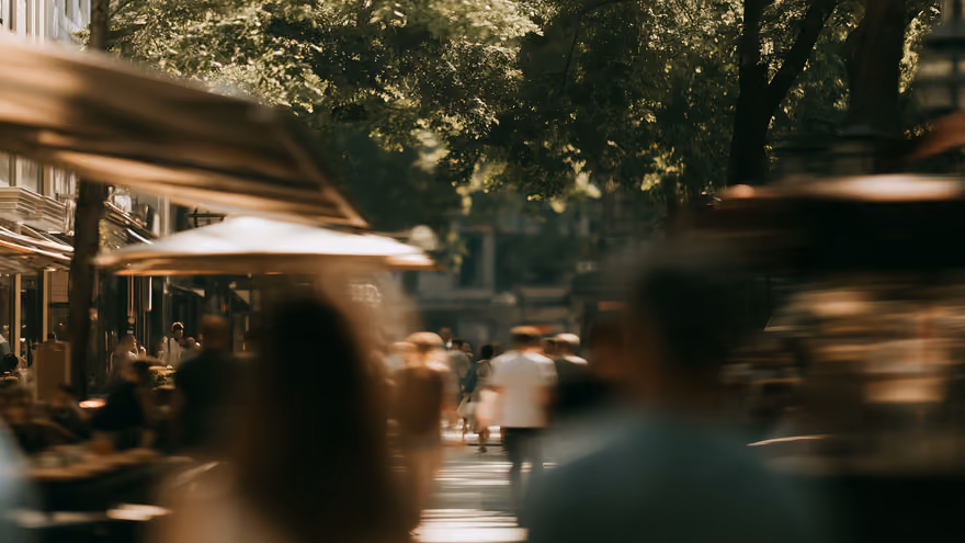 A busy city street in summer, pedestrians blurred by motion under trees.